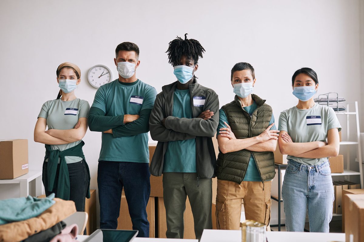 Diverse team of volunteers wearing masks while standing with arms crossed during help and donation event, copy space